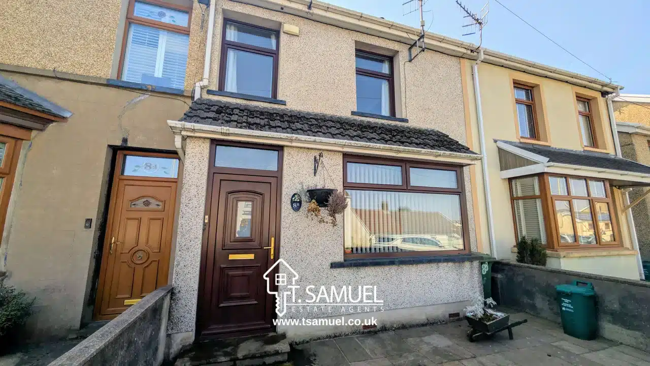 Front view of a two-story terraced house with a brown front door, large front window, and a hanging plant basket; estate agent sign visible on the window.