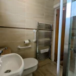 Small beige-tiled bathroom with a toilet, sink, heated towel rail, and a glass shower enclosure. Grab bars are installed near the toilet. Branding and website text appear in the lower left corner.
