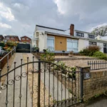 A detached house with a gravel driveway, gated entrance, and small front garden, located on a slightly elevated plot in a suburban neighborhood.