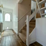 View of a bright hallway with wooden flooring, a staircase with carpeted steps, a white front door, and a glimpse of a kitchen on the left. Estate agent branding is visible on the image.