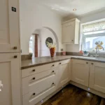 Bright kitchen with white cabinets, stone countertops, stainless steel sink, and a window. A vase with flowers sits on the counter, and natural light fills the room.