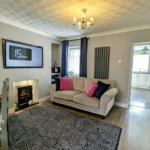 Modern living room with a beige sofa, patterned rug, wall-mounted TV, electric fireplace, dining area, and view into a white kitchen.