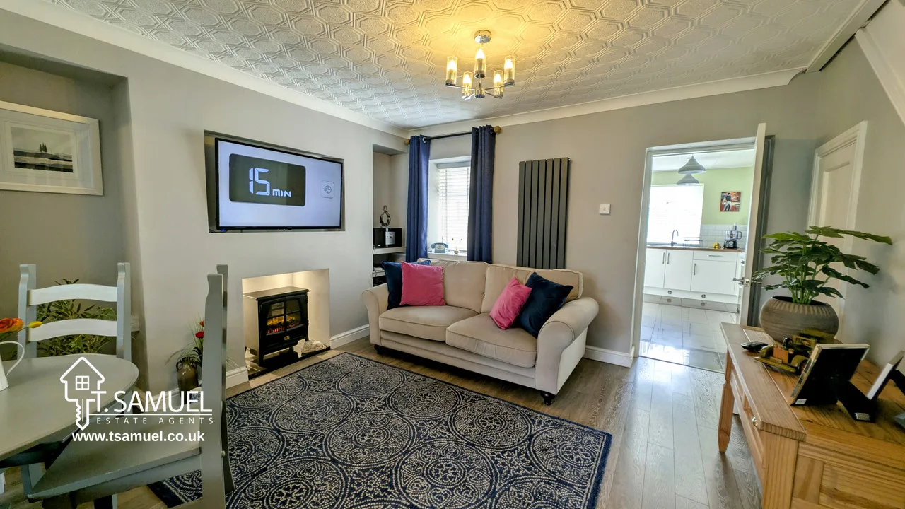 Modern living room with a beige sofa, patterned rug, wall-mounted TV, electric fireplace, dining area, and view into a white kitchen.
