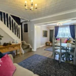 Open-plan living and dining area with a staircase, wooden floors, blue rugs, and a dining table; bright natural light enters through large window with curtains.