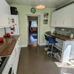 Modern galley kitchen with white cabinets, wooden countertops, barstools, and patterned wallpaper; doorway leads to living area with a pink sofa. T. Samuel Estate Agents logo in corner.