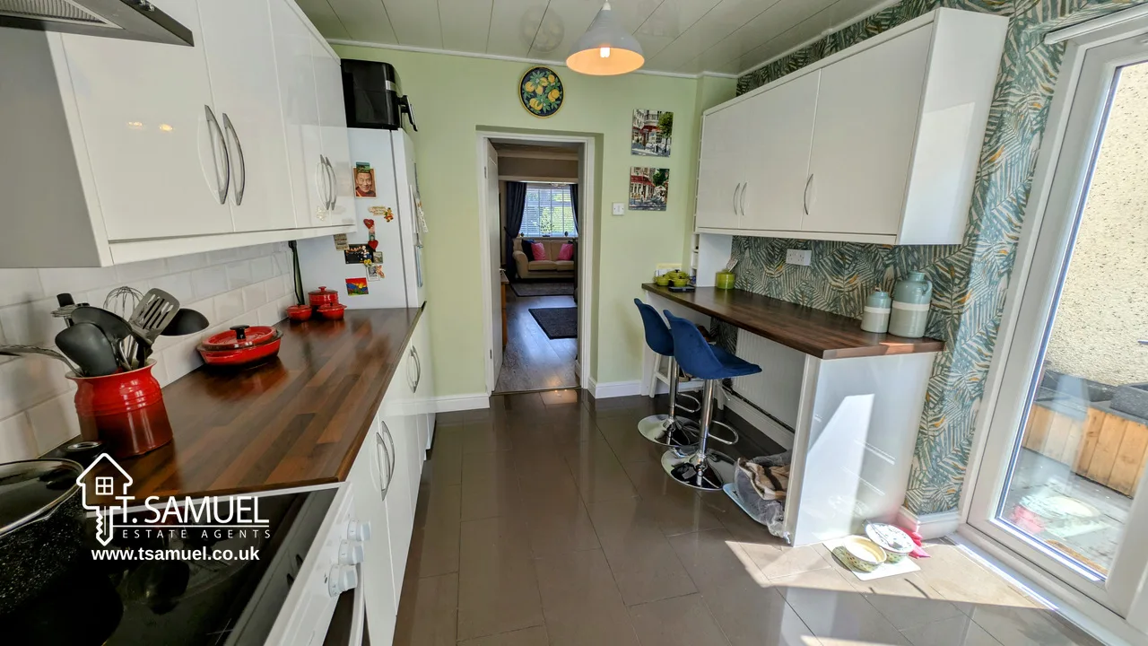Modern galley kitchen with white cabinets, wooden countertops, barstools, and patterned wallpaper; doorway leads to living area with a pink sofa. T. Samuel Estate Agents logo in corner.
