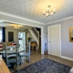 A modern living and dining area with grey walls, wooden flooring, a dining table, staircase, chandelier, and decorative items including a Buddha statue and framed mirror.