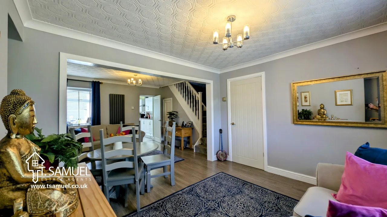 A modern living and dining area with grey walls, wooden flooring, a dining table, staircase, chandelier, and decorative items including a Buddha statue and framed mirror.