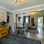 A modern living and dining area with wooden floors, grey walls, a dining table, couches, decorative items, and large rugs. Natural light enters from windows in the background.