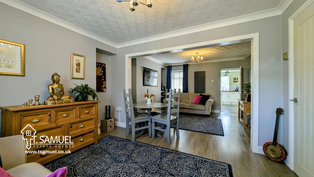 A modern living and dining area with wooden floors, grey walls, a dining table, couches, decorative items, and large rugs. Natural light enters from windows in the background.