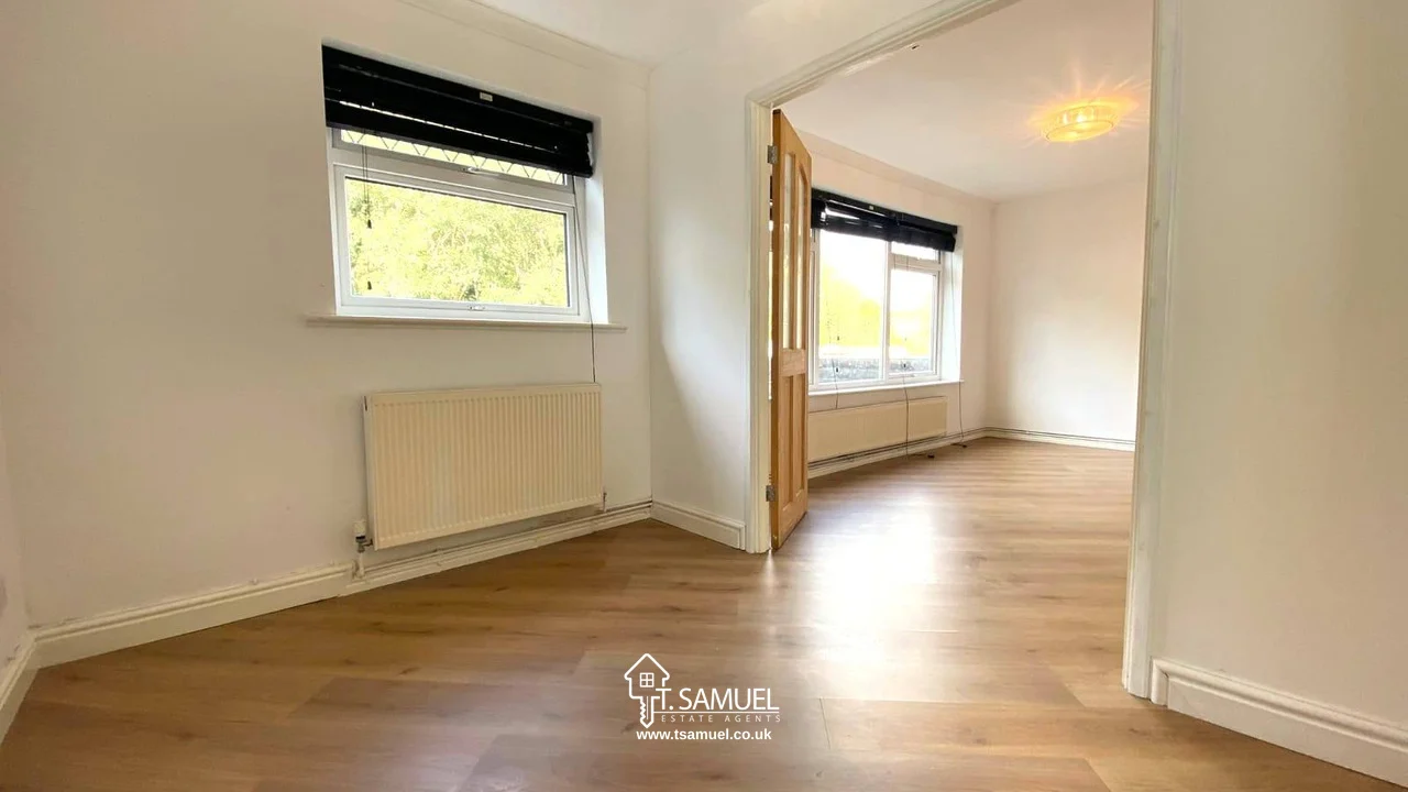 Bright, empty room with light wood flooring, white walls, large windows with black blinds, and a radiator beneath one window. Glass double doors lead to an adjacent room.
