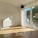 A bright, empty room with wooden flooring, a white radiator, and large glass sliding doors opening to a small patio and green outdoor area.
