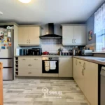Modern kitchen with light cabinets, black appliances, a dining table with chairs, a fridge covered in magnets, and a window with white lace curtains letting in natural light.