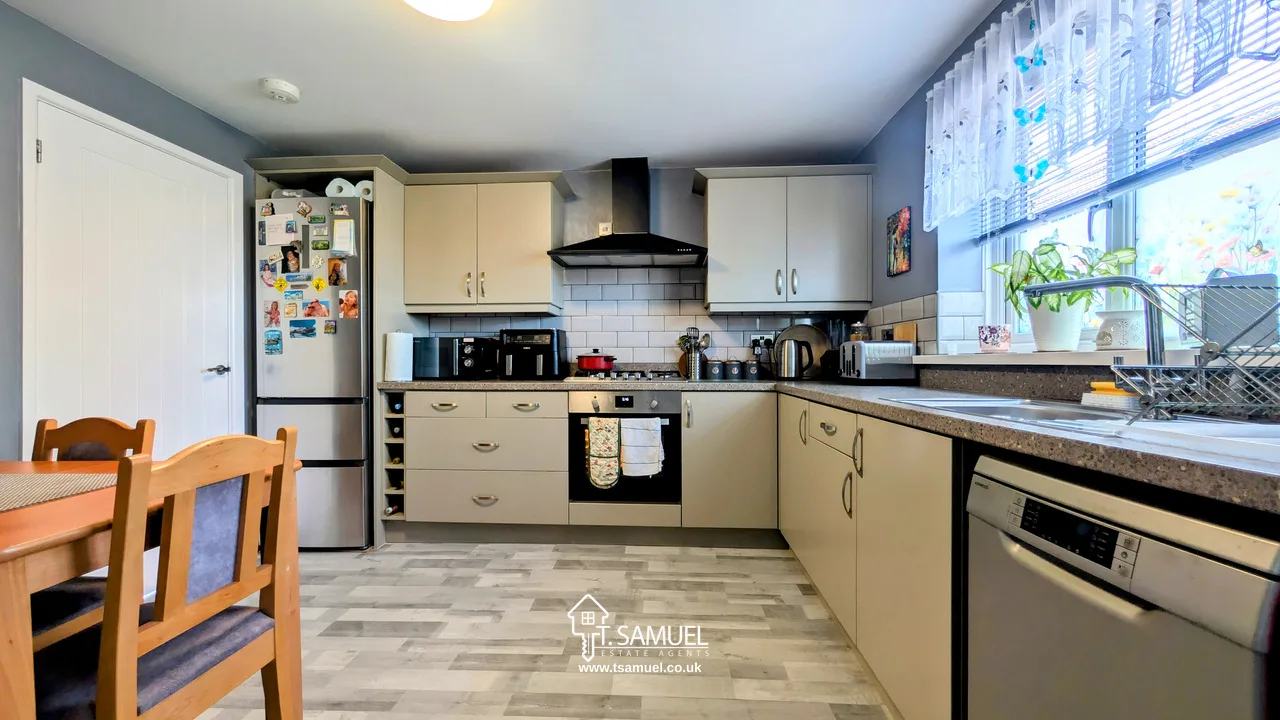 Modern kitchen with light cabinets, black appliances, a dining table with chairs, a fridge covered in magnets, and a window with white lace curtains letting in natural light.