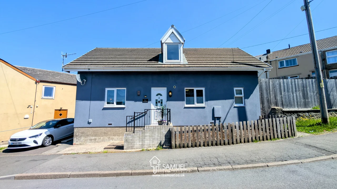 A blue detached house with a dormer window, white door, small fenced yard, and a car parked on the street in front.