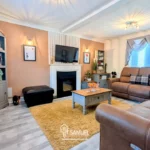 Well-lit living room with brown leather sofas, a coffee table, a fireplace beneath a wall-mounted TV, bookshelves, and window with drapes. Neutral and pastel colors dominate the decor.