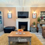 A cozy living room with a TV above a fireplace, flanked by cabinets and shelves, a brown armchair, a coffee table with fruit, and a tan rug on a light gray floor.