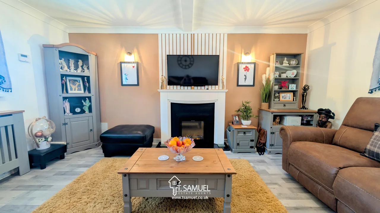 A cozy living room with a TV above a fireplace, flanked by cabinets and shelves, a brown armchair, a coffee table with fruit, and a tan rug on a light gray floor.