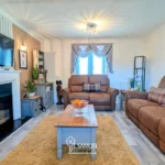 A cozy living room with brown sofas, a coffee table, a wall-mounted TV above a fireplace, and a window with drapes letting in natural light.