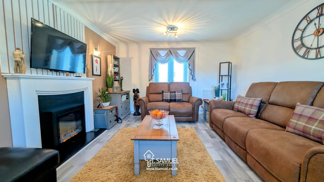 A cozy living room with brown sofas, a coffee table, a wall-mounted TV above a fireplace, and a window with drapes letting in natural light.
