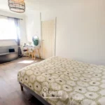 Bright bedroom with a double bed, small vanity table, radiator, and window with sheer curtains. The room has wood flooring and minimal decor.