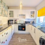 Modern kitchen with white cabinets, black countertops, oven, microwave, toaster, washing machine, and bright yellow window blind. A drying rack and various appliances are on the counters.