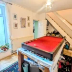 A room with a red pool table in the center, plants by a window, wall decor, a staircase, and various household items scattered around.