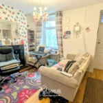 A living room with patterned wallpaper, sofa, armchair, TV, small desk, rug, and chandelier, featuring personal decor and a front door visible in the background.