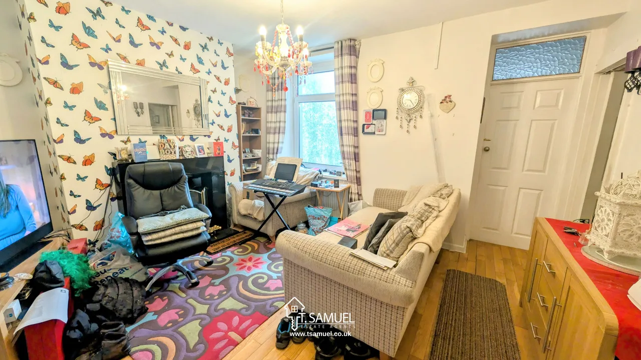 A living room with patterned wallpaper, sofa, armchair, TV, small desk, rug, and chandelier, featuring personal decor and a front door visible in the background.