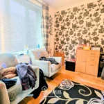 A small room with two armchairs, a patterned black and white wall, a window with blinds and curtains, a cabinet with books, and various items scattered around.