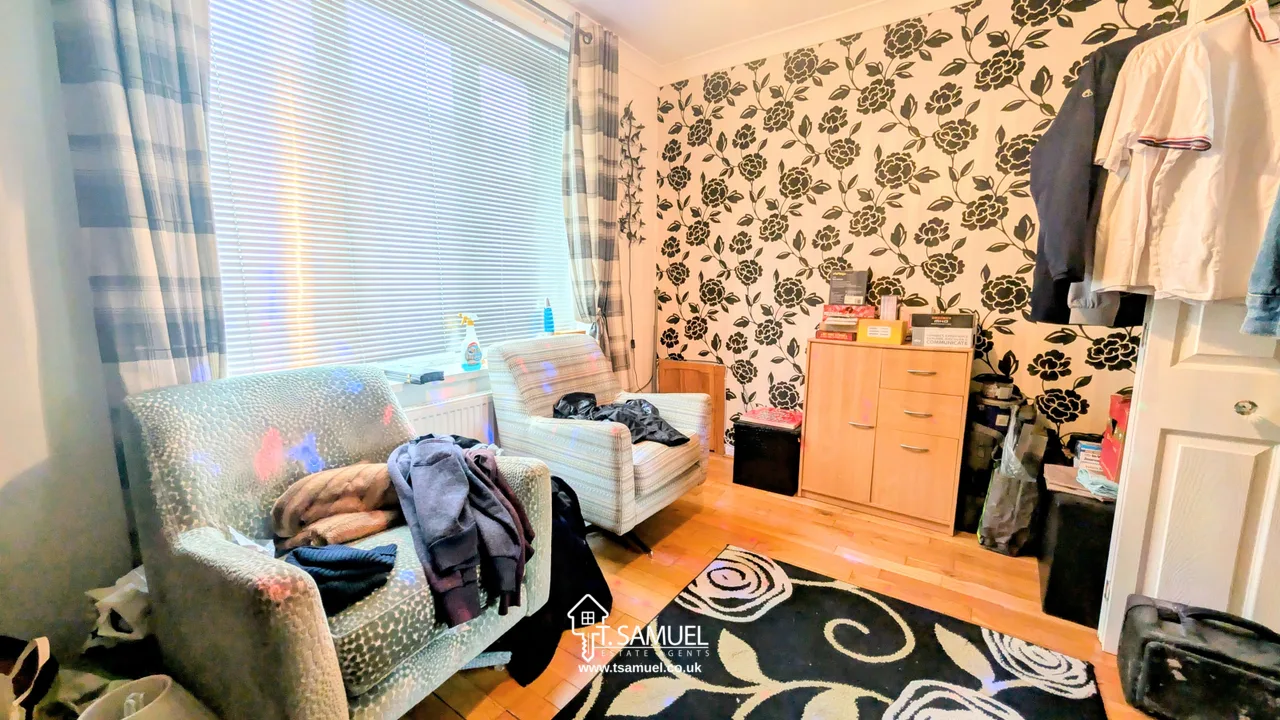 A small room with two armchairs, a patterned black and white wall, a window with blinds and curtains, a cabinet with books, and various items scattered around.