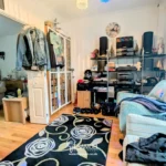 A living room with armchairs, a patterned rug, shelves with electronics and decor, jackets hanging by a doorway, and a view into another room with a sofa.