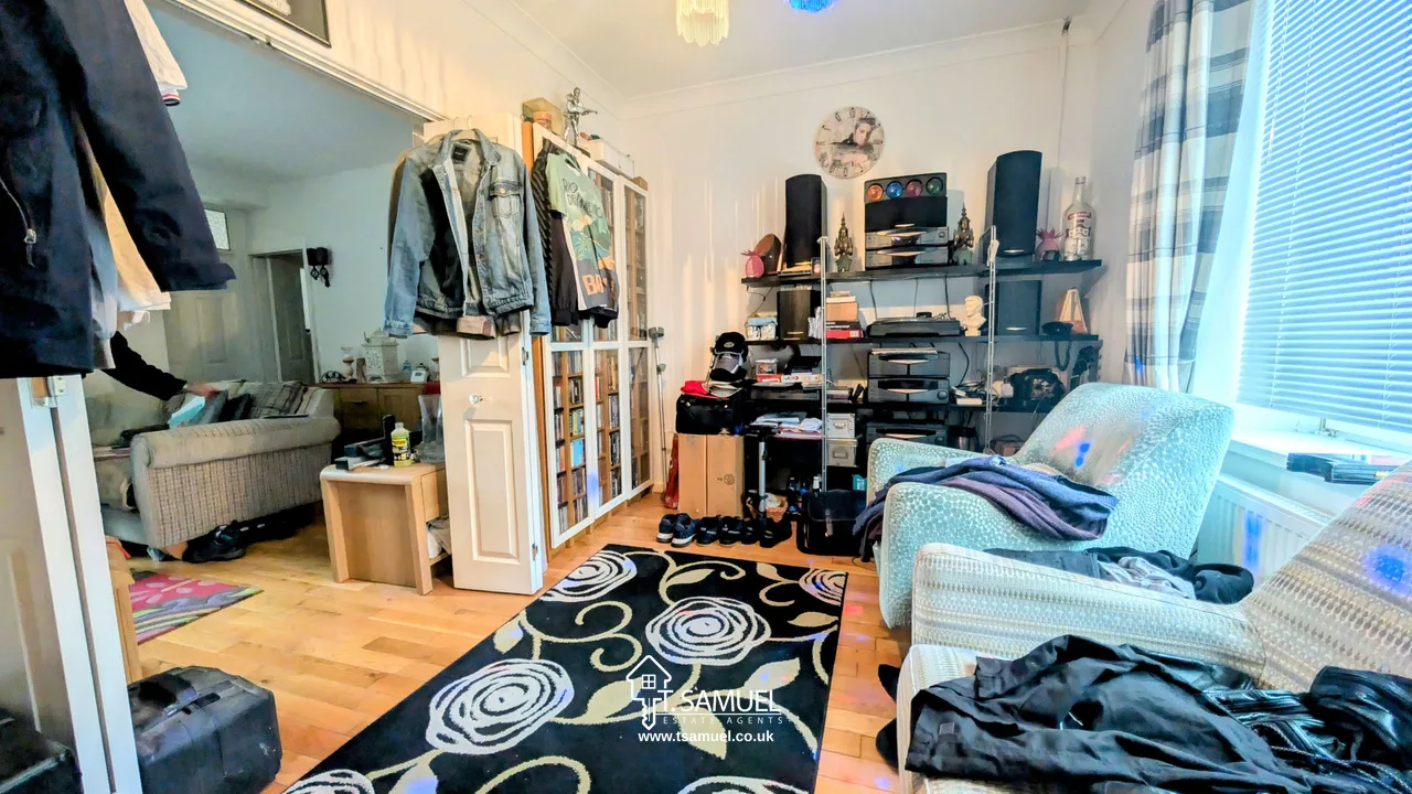 A living room with armchairs, a patterned rug, shelves with electronics and decor, jackets hanging by a doorway, and a view into another room with a sofa.