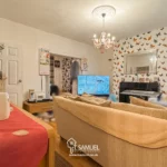 A living room with a beige sofa, TV, piano, butterfly-patterned wallpaper, chandelier, and shelves. Red table runner with decor in the foreground. The photo has a watermark for "T. Samuel".