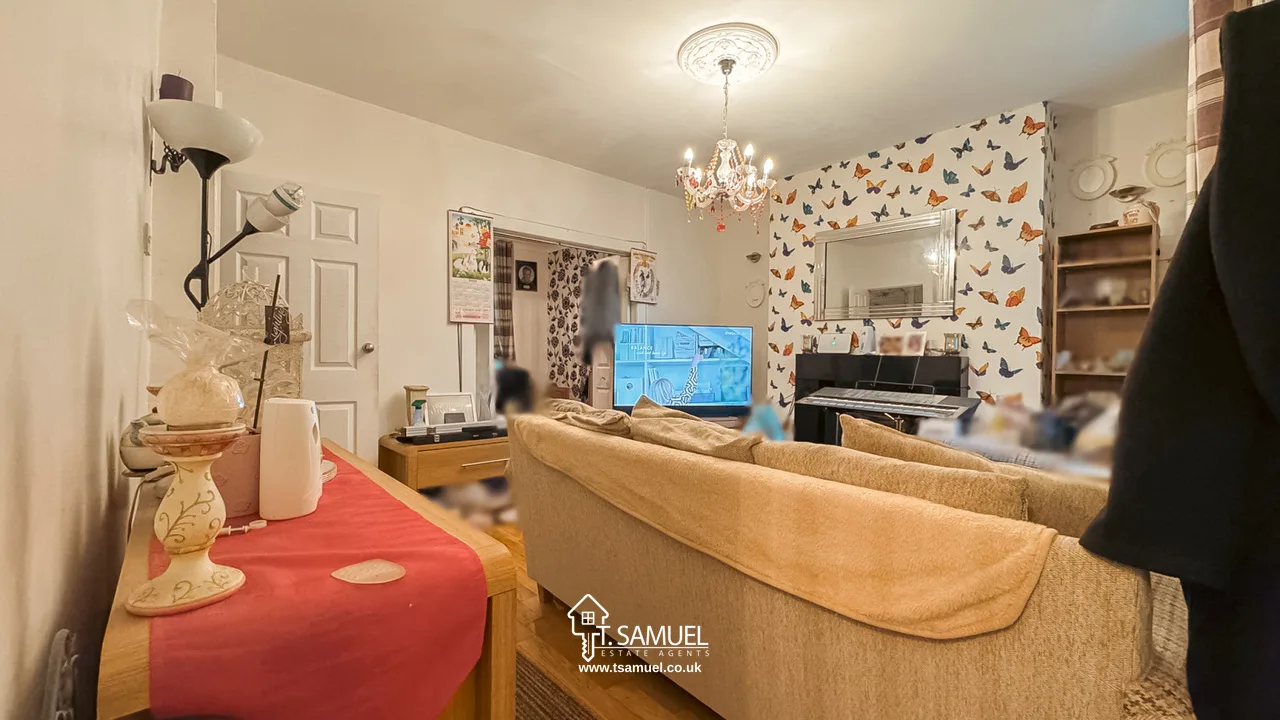 A living room with a beige sofa, TV, piano, butterfly-patterned wallpaper, chandelier, and shelves. Red table runner with decor in the foreground. The photo has a watermark for "T. Samuel".