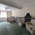A living room with patterned wallpaper, green carpet, armchairs, a sofa, fireplace, window with curtains, and cardboard boxes on the floor. T. Samuel Estate Agents logo is visible.