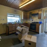 A modest kitchen with wooden cabinets, scattered cleaning supplies on the counter, open moving boxes on the floor, and a back door letting in light. T. Samuel Estate Agents watermark present.