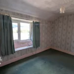 Empty bedroom with patterned wallpaper, green carpet, a window with curtains, a ceiling light, and a closed white door. Estate agent logo and web address in bottom left corner.
