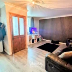 A living room with wood flooring, a brown leather sofa, wall-mounted shelves, a TV on a white unit, and a wooden door; coats hang on the left near a window.