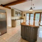 Spacious modern kitchen with island, granite countertops, stainless steel fridge, cream cabinets, large windows, and tiled floor. Estate agent logo in lower left corner.