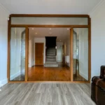 A view from a room with light wood flooring through glass sliding doors into a hallway with wooden floors, a staircase, and a wooden door at the far end. Vintage trunks are on the right.