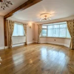 Spacious, empty living room with wooden floors, bay window, cream walls, ceiling lights, and floral curtains. Estate agent logo in lower left corner.
