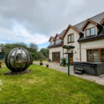 A large house with multiple dormer windows, a garden lawn, a modern spherical garden pod, a bird feeder, and a hot tub on a stone patio.