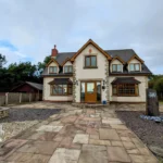 A two-story house with cream walls, brown trim, and a stone pathway in front, surrounded by trees and a gravel yard. The T. Samuel estate agency logo is visible in the lower left corner.