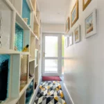 A narrow hallway with patterned carpet, glass shelving on the left, framed pictures on the right, and a glass door at the end letting in natural light.