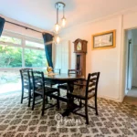 A dining room with a wooden table and six chairs, a large window with blue curtains, a grandfather clock, patterned carpet, and framed wall art.
