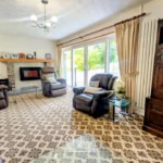 Living room with patterned carpet, three recliner chairs, a fireplace, wooden cabinet, glass coffee table, and large windows with beige curtains showing greenery outside.